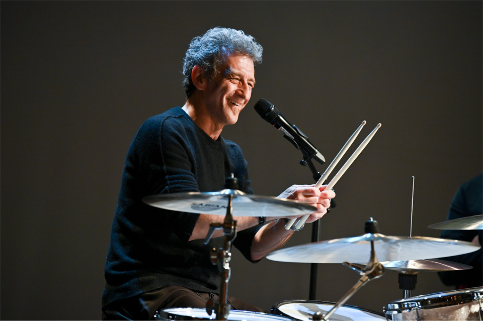 Man, wearing a dark top, smiling at the audience, holding a pair of drum sticks, sitting at a drum kit.